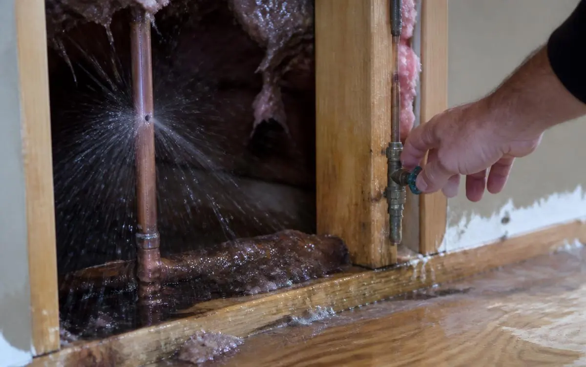 Water spraying from a burst copper water pipe inside an opened wall cavity while a hand reaches for the shutoff valve